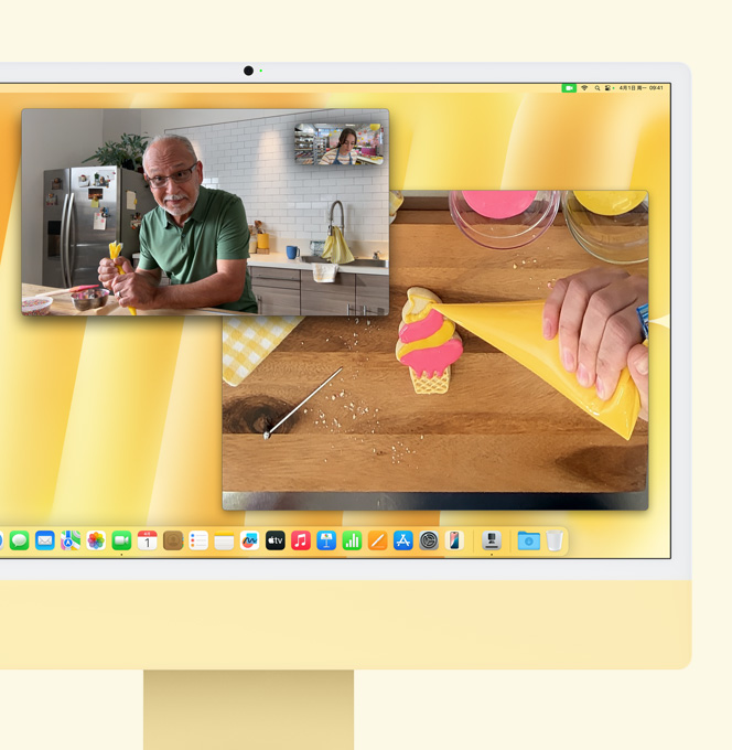 黄色 iMac，演示桌上视角功能。屏幕显示烘焙师正在与另一个人视频通话。一个屏幕显示烘焙师和男子的脸。另一个屏幕显示烘焙师的手在为曲奇饼干添加糖花。