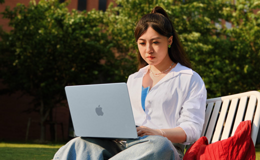 照片展示一位女生坐在草坪边的长椅上用着 MacBook Air。