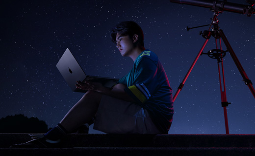 照片展示星空下，一位男生坐在天文望远镜前用着 MacBook Air。