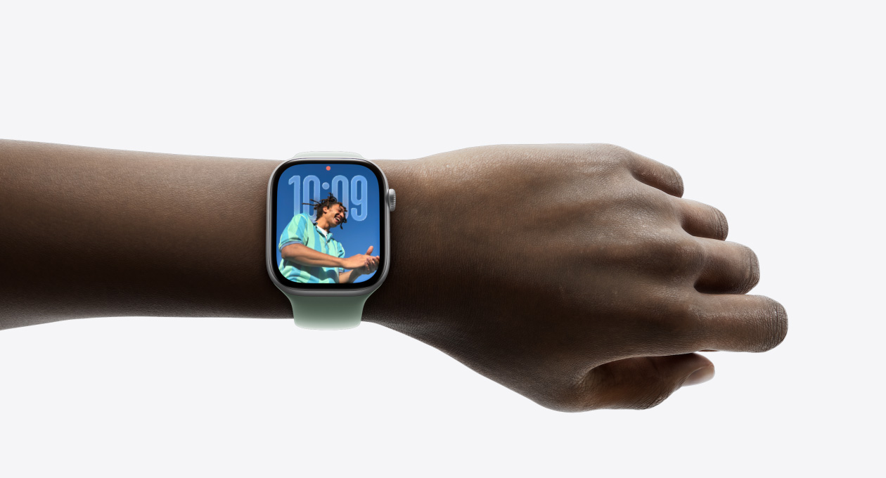 特写画面显示一只手腕戴着配有橡胶表带的银色 Apple Watch，表盘展示全新 Liquid Glass 设计的时间 app。