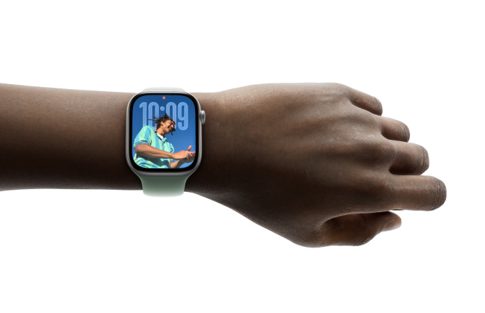 特写画面显示一只手腕戴着配有橡胶表带的银色 Apple Watch，表盘展示全新 Liquid Glass 设计的时间 app。